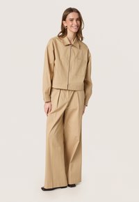 Beige cropped jakke med lynlås foran, sidelommer og matchende brede bukser, begge i glat stof. Modellen står foran en ensfarvet baggrund.