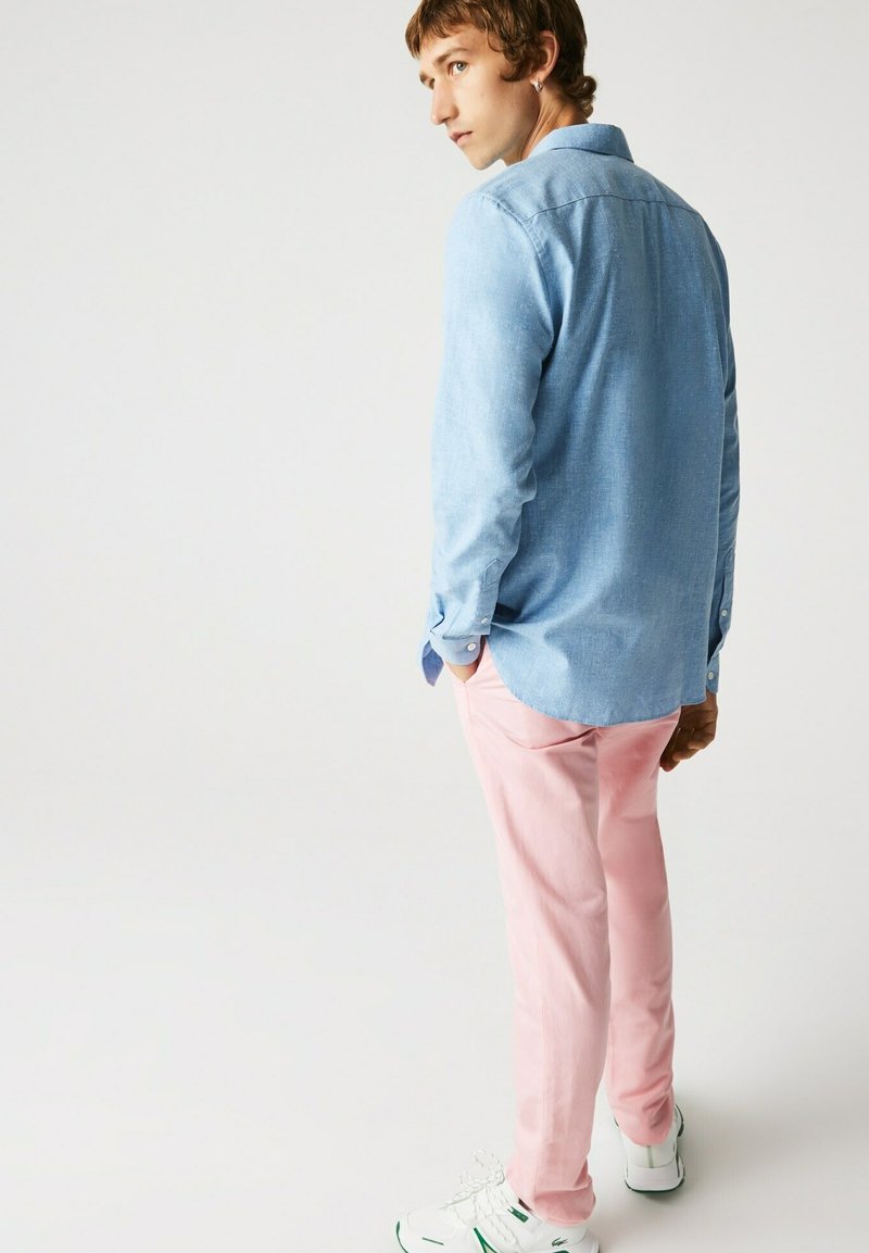 Himmelblaues Hemd mit Knöpfen, pinkfarbene Hose, weiße Sneakers mit grünen Akzenten. Das Hemd hat einen entspannten Schnitt und sichtbare Nähte.