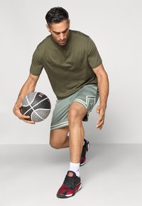 Αθλητική εμφάνιση με σκούρο πράσινο κοντομάνικο μπλουζάκι και ανοιχτό πράσινο σορτσάκι με λευκές λεπτομέρειες; μαύρη μπάλα μπάσκετ και μαύρα-κόκκινα αθλητικά παπούτσια.