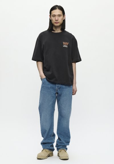Tricou negru oversize cu un logo grafic pe piept, asortat cu blugi albaștri lejeri și pantofi slip-on bej. Textură fină de bumbac.