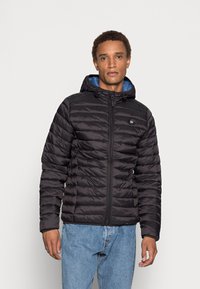 Svart puffjakke med horisontalt kviltet mønster, glidelås lukking og blå hettefôr. Brukt over lyseblå jeans.