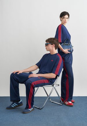 Άνδρας που κάθεται σε καρέκλα και γυναίκα όρθια, και οι δύο φορούν ταιριαστά σκούρα μπλε αθλητικά κοστούμια με κόκκινες ρίγες, αθλητικά παπούτσια, και η γυναίκα κρατάει μια μπλε τσάντα.