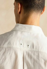 Gaišu krāsu krekls ar skatu no mugurpuses, klasiskā apkakle, divas caurumi un plisējums zem kakla, lai pievienotu dizaina detaļas.