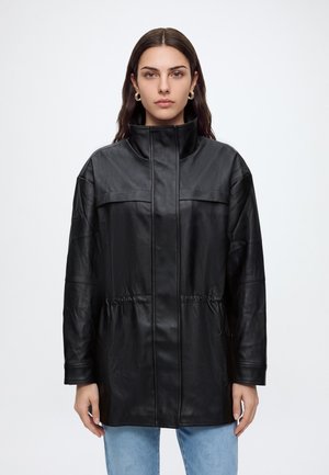 Jeune femme aux longs cheveux foncés portant des boucles d'oreilles créoles dorées, une veste en cuir noire à col haut et un jean bleu clair sur un fond uni.