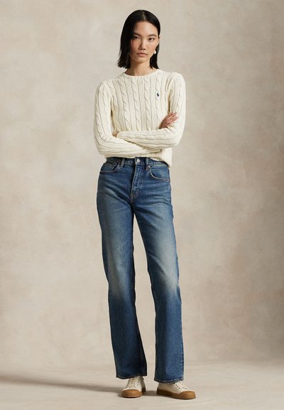 Femme debout, les bras croisés, portant un pull torsadé crème, un jean bleu taille haute et des baskets beiges sur un fond uni.