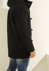Parka negra con capucha, con cierres de botones y bolsillos laterales, hecha de una tela mate. Llevada sobre unos jeans de color azul claro.