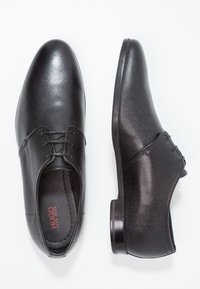 Chaussures en cuir noir à finition texturée, dotées de lacets et d'un bout arrondi. La semelle est lisse et légèrement surélevée, avec des coutures contrastantes.