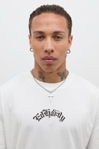 Biele bavlnené tričko s čiernym logom "Ed Hardy", nosené so strieborným krížom a kruhovými náušnicami, s tetovaniami na krku.
