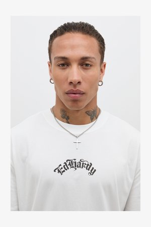 Biała bawełniana koszulka z czarnym logo "Ed Hardy", noszona z srebrnym naszyjnikiem z krzyżem i kolczykiem w kształcie kółka, z tatuażami na szyi.