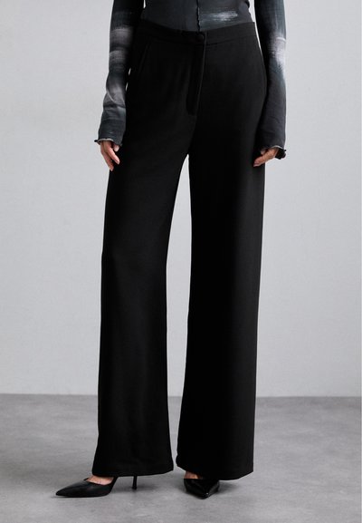 Pantaloni neri a gamba larga con texture liscia, dotati di chiusura con bottone e tasche frontali, abbinati a tacchi a spillo neri.