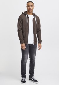 Brun zip-up hoodie med en hvid snor, iført over en hvid t-shirt. Kåret med mørkegrå slim-fit jeans og sorte sneakers.