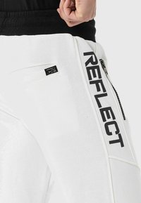Weiße Jogginghosen mit schwarzen Akzenten, featuring eine Reißverschlusstasche und dem Aufdruck "REFLECT" entlang der Seite in fetten schwarzen Buchstaben.