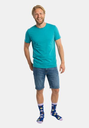 Türkisfarbenes T-Shirt mit kurzen Ärmeln, Jeansshorts und blaue Socken mit Keks-Muster. Das Modell steht lächelnd mit den Händen in der Hüfte.