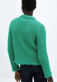 Turkusowy kardigan o fakturowanej powierzchni z tkanym wzorem, kołnierzem i surowym wykończeniem na dole, zestawiony z ciemnymi dżinsami. Materiał z mieszanki wełny.