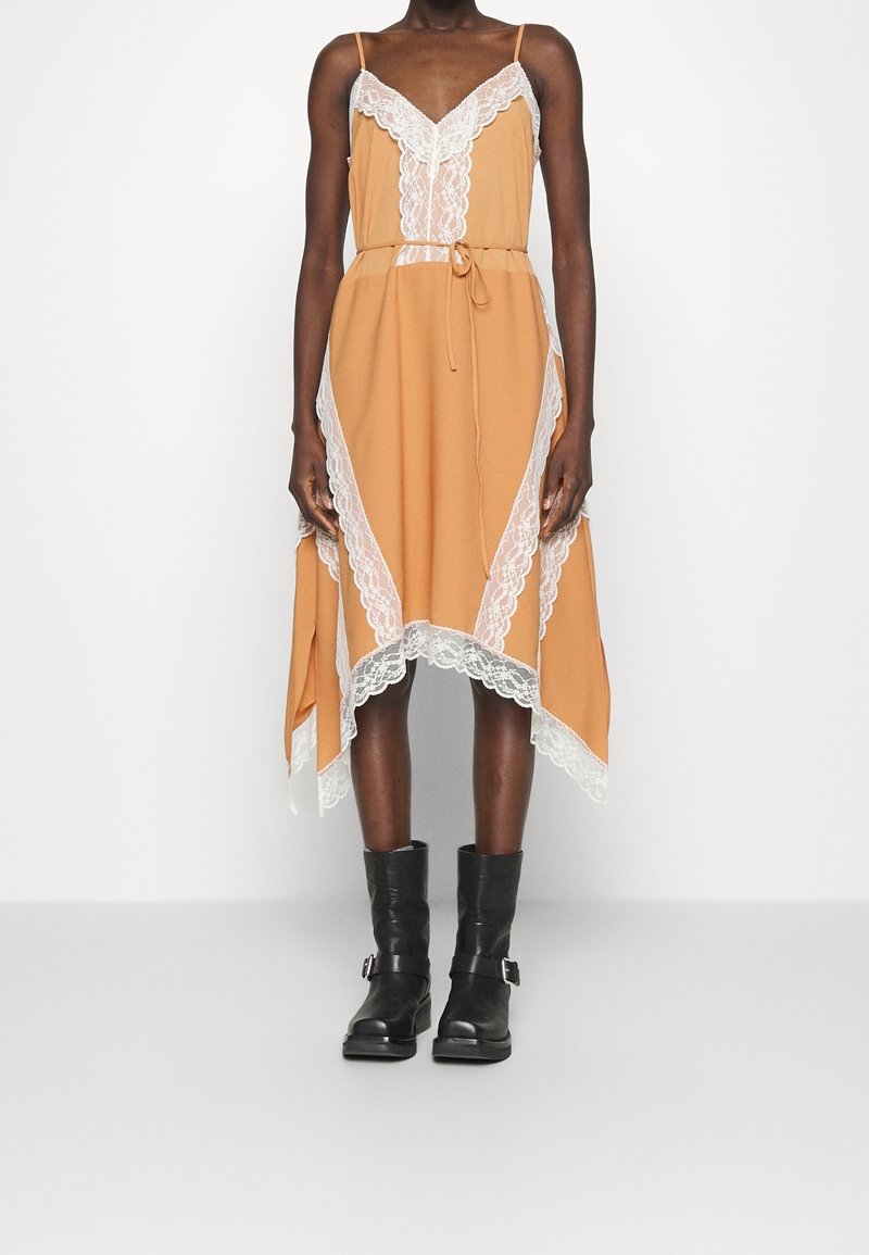 Femme portant une robe nuisette beige à bordure en dentelle blanche et taille nouée, associée à des bottes en cuir noir mi-mollet avec des boucles.