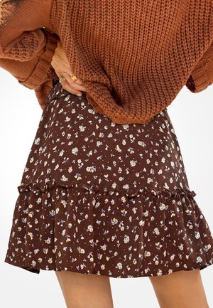Kobieta w brązowym swetrze dzianinowym i brązowej spódnicy w kwiaty z falbanami, z ręką opartą na biodrze, sfotografowana od połowy uda do talii.