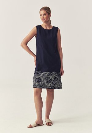 Femme debout portant une robe sans manches bleu marine avec des motifs de feuilles blanches près de l'ourlet et des sandales beige à enfiler sur un fond uni.