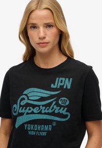 Sort bomulds T-shirt med turkis grafisk tryk med teksten "Superdry," "JPN," og "Yokohama 'High Flyers.'" Rund halsudskæring, korte ærmer.