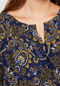 Blusa floreale blu navy con motivi paisley gialli e bianchi, scollo a V con chiusura a bottone e una texture di tessuto leggera.