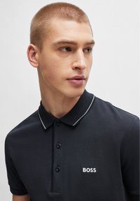 Svart polo-skjorte med teksturert stoff, hvit knappestolpe og kontrasterende hvit kragekant. Har brodert "BOSS" logo i hvitt.
