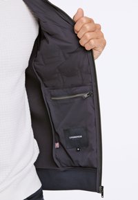 Svart jacka med en slät inredning, med en dragkedjeficka och en etikett som säger "LINDBERGH", storlek medium längst ner.