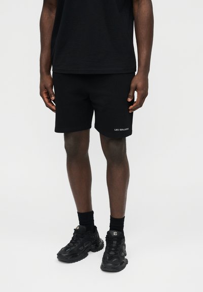 Άνδρας με μαύρο σορτς Les Benjamins, μαύρο μπλουζάκι, μαύρες κάλτσες και μαύρα chunky sneakers, στέκεται μπροστά σε λευκό φόντο.