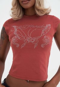 Korte mouwen cropped T-shirt in roestkleur, met witte strassversieringen in een bloemmotief over de voorkant en een ronde halslijn.
