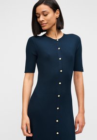 Φόρεμα ναυτικού μπλε με ριμπ ύφανση, κοντά μανίκια, στρογγυλή λαιμόκοψη και χρυσά κουμπιά στο μπροστινό μέρος. Το εφαρμοστό σχέδιο αναδεικνύει τη σιλουέτα.