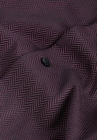 Schwarzer Knopf auf violettem Stoff mit diagonalem Fischgrätenmuster, das Textur und Falten zeigt.