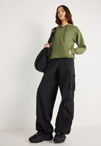 Olivovo zelená mikina s kapucňou, predným vreckom a šnúrkou, kombinovaná s čiernymi nákladnými nohavicami s viacerými vreckami a zipsami, doplnená čiernymi teniskami.