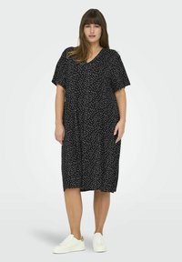 Rochie neagră cu decolteu în V, mâneci scurte și croială lejeră. Are un model cu buline albe. Este asortată cu adidași albi pentru contrast.