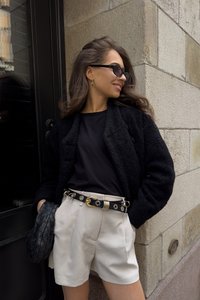 Musta teksturoitu takki, musta t-paita, kermaiset shortsit poimutuksilla, musta nahkavyö kultaisilla yksityiskohdilla ja musta kudottu clutch-laukku.