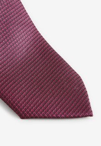 Sügava punase ja lilla mustriga textuuriga lipsu ots valgel taustal lähivõttepildis.