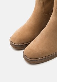 Brązowe zamszowe botki za kostkę z okrągłym noskiem, z zauważalnym detalem szycia wzdłuż podeszwy oraz miękką fakturą.