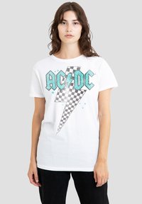 Junge Frau mit welligem braunem Haar, die ein weißes AC/DC Grafik-T-Shirt mit einem schwarz-weißen karierter Blitz trägt, dazu schwarze Hosen.