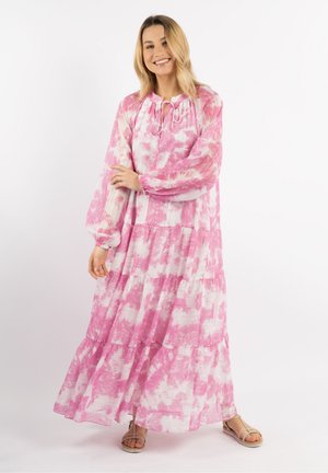 Frau lächelt, trägt ein langes rosa-weißes Batikkleid mit langen Ärmeln und beigen Sandalen, steht vor einem weißen Hintergrund.