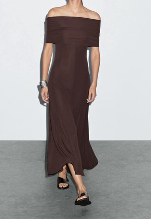 Donna che cammina con un vestito marrone scuro lungo fino ai piedi, con spalle scoperte, indossando un braccialetto d'argento e sandali neri bassi.