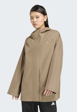 Vrouw draagt een losse beige capuchonjas en een zwarte broek met een wit logo, staand tegen een effen lichte achtergrond.