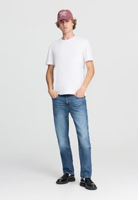 Giovane uomo che indossa una maglietta bianca, jeans blu, scarpe nere e un cappellino bordeaux, in piedi con le mani nelle tasche su uno sfondo neutro.