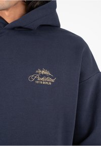 Sudadera azul marino de algodón suave con un ajuste relajado. Presenta un logo bordado en dorado y el texto: "Prohibido 10119 BERLÍN" en la parte delantera.