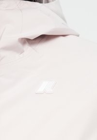 Cazadora de color rosa claro con cuello alto, que presenta un logo blanco brillante en el lado izquierdo. La tela parece suave y ligera.