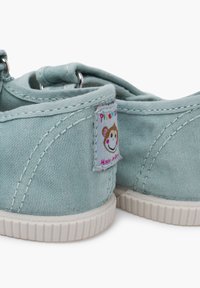 Zapatillas de lona azul claro con superficie texturizada, suelas de goma blancas y detalle de costura. Presenta una etiqueta con un diseño de mono sonriente.