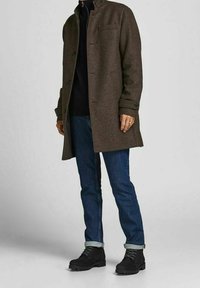 Personne portant un manteau marron boutonné, un col roulé noir, un jean bleu retroussé et des bottes noires à lacets, debout devant un fond uni.