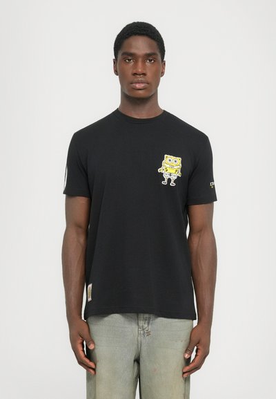 Man wearing black T-shirt with small yellow SpongeBob graphic on chest and light faded jeans, standing against plain white background.