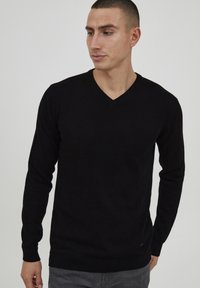 Maglione nero a V in morbido punto maglia, con maniche lunghe e orlo a coste, indossato con pantaloni grigi.
