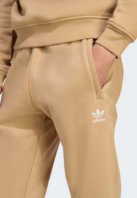 Beżowe dresy z bawełny z elastycznym pasem, bocznymi kieszeniami i białym logo Adidas na lewym biodrze. Gładka faktura, luźny krój.