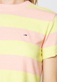 Acercamiento a una camiseta de rayas en tonos rosa pastel y amarillo, con mangas cortas y un pequeño logotipo rectangular en rojo, blanco y azul en el pecho.
