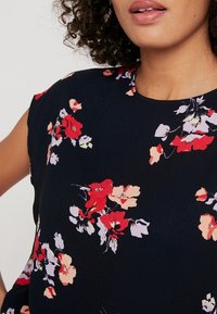 Blommig blus i mörk marinblått tyg, med flerfärgade blommönster i rött, rosa och vitt. Slät yta med rund halsringning.