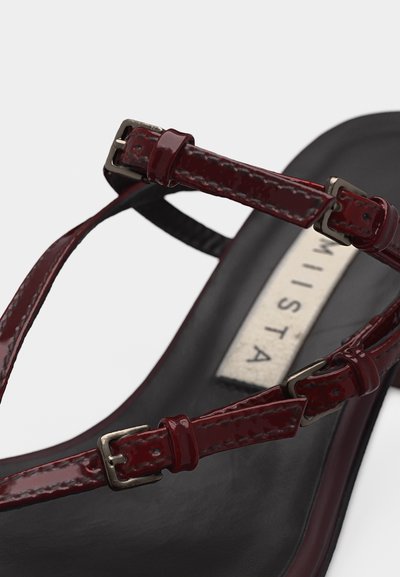 Μαύρο σανδάλι με σκούρες κόκκινες γυαλιστερές λουριά και ασημένιες αγκράφες, με ορατή ετικέτα στην πατούσα.
