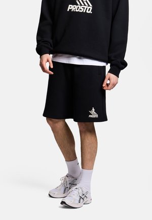 Osoba ubrana w czarne spodenki Prosto, czarną bluzę Prosto, białe skarpetki oraz białe buty sportowe Asics, stojąca na jednolitym tle.